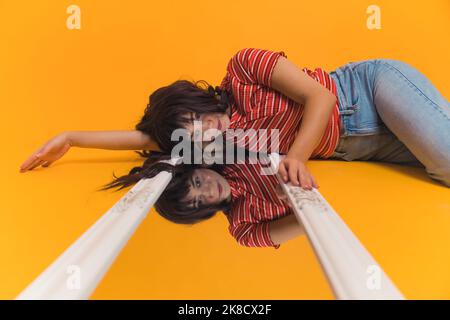Young, bright, caucasian girl with long brown hair laying on the mirror on the floor. Girls reflection in the mirror, isolated orange background. High quality photo Stock Photo