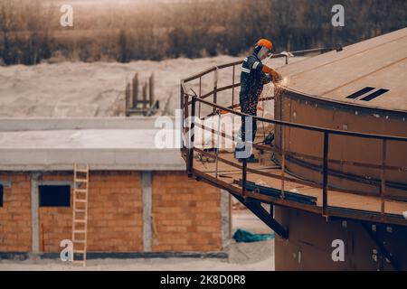 Worker in protective uniform and helmet grinding top of tower on construction site. Stock Photo