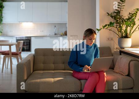 Young freelancer sitting on cozy sofa works on laptop and makes business project in apartment Stock Photo