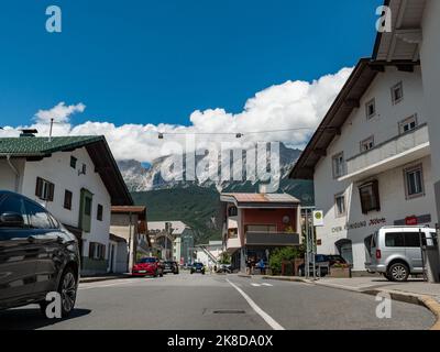 Street view in Tirol. People and cars are moving in the town. The road is going up steep and mountains of the Alps are in the background. White clouds Stock Photo