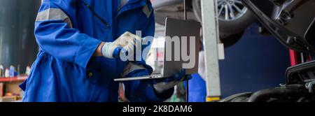 Young man is mechanic using laptop computer checking engine of car in the garage, worker examining and diagnostic automobile, technician and maintenan Stock Photo