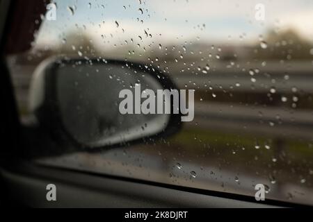 Rear-view mirror of car behind wet glass. View of road in rain. Details of trip. Drops on glass. Stock Photo