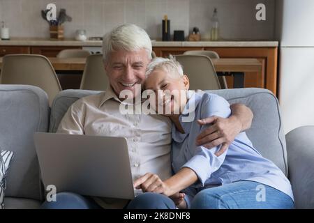 Happy older spouses relaxing on sofa at home using laptop Stock Photo