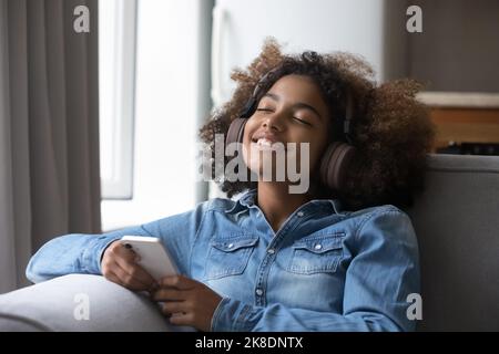 Peaceful African teenage girl holds cellphone listens music through headphones Stock Photo