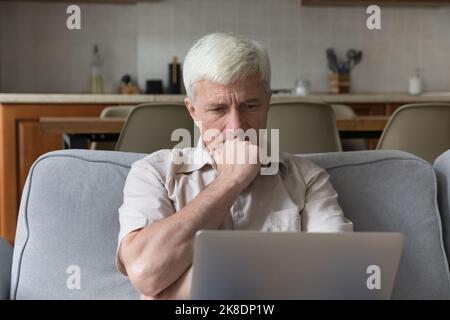 Serious thoughtful older man sit on couch with laptop Stock Photo