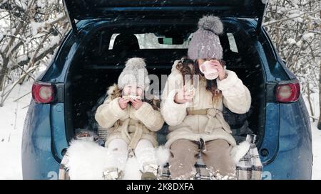 winter tea picnic. Happy cutie, little girl, dressed in warm winter clothes, are having tasty snack, tea party outdoors. They are sitting on car trunk, in snowy forest, during snowfall. winter family fun. happy time on snowy winter day. winter family activity outdoors. High quality photo Stock Photo