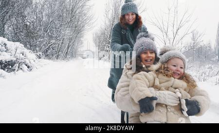 family sledding in winter. outdoor winter activity. Happy, laughing family, woman with 2 daughters are enjoying of sledging on snowy road, in forest, during snowfall. family is having fun, spending time together on snowy winter day. slow motion. High quality photo Stock Photo