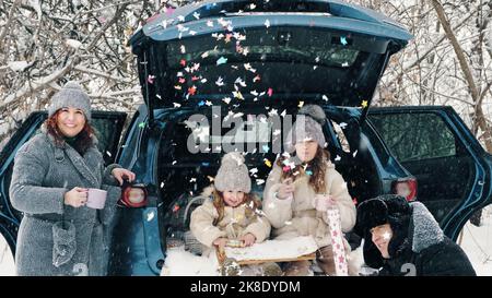 winter tea picnic. Happy family is having tasty snack, a tea party with cream, outdoors. They are sitting on car trunk, in snowy forest, during snowfall. winter family fun. happy time on snowy winter day. winter family activity outdoors. High quality photo Stock Photo