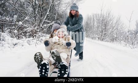 family sledding in winter. outdoor winter activity. Happy, laughing family, woman with 2 daughters are enjoying of sledging on snowy road, in forest, during snowfall. family is having fun, spending time together on snowy winter day. slow motion. High quality photo Stock Photo