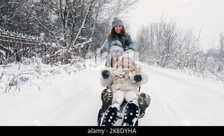 family sledding in winter. outdoor winter activity. Happy, laughing family, woman with 2 daughters are enjoying of sledging on snowy road, in forest, during snowfall. family is having fun, spending time together on snowy winter day. slow motion. High quality photo Stock Photo