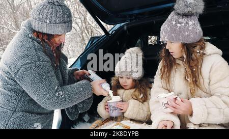 winter tea picnic. Happy family is having tasty snack, a tea party with cream, outdoors. They are sitting on car trunk, in snowy forest, during snowfall. winter family fun. happy time on snowy winter day. winter family activity outdoors. High quality photo Stock Photo