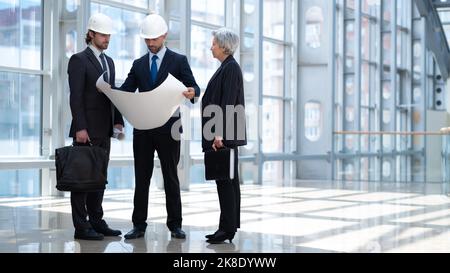 Multi ethnic business architect people in safety helmets discussing blueprint standing in the lobby at office Stock Photo