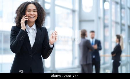 Confident businesswoman talking on a phone, female team leader posing for photo, standing in modern office with diverse team on background Stock Photo
