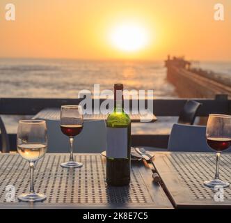 Bottle of Red Wine with three glasses on a table, with a beautiful sunset background - Swakopmund Pier, Namibia Stock Photo