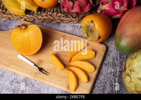 Persimmon cut into pieces on a wooden board, accompanied by tropical fruits such as pitahaya, mango, banana and custard apple. Stock Photo