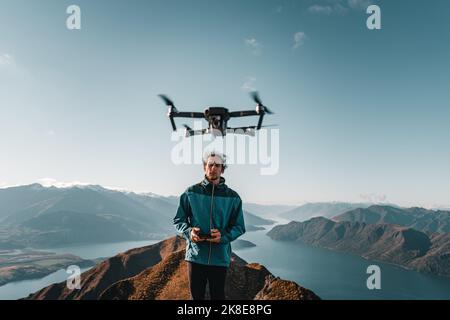 young caucasian man with blue jacket disheveled black pants looking at camera and operating the remote control of a drone that is flying in the air Stock Photo