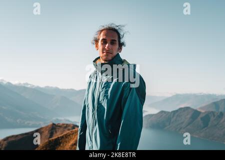 disheveled caucasian boy in blue coat looking at camera calm and smiling on quiet lonely mountain summit above big blue water lake, roys peak, new Stock Photo