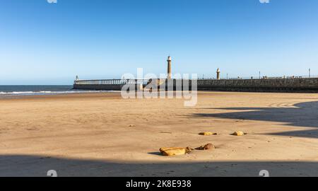 A pier with a lighthouse stretches out to sea. Rocks and shadows are on the sandy beach and a clear sky is above. Stock Photo