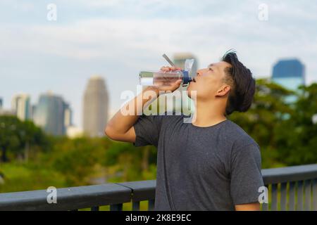 Image of Asian healthy man holding a bottle of refreshing water, resting after a workout or a run in city park Stock Photo