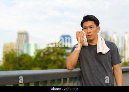 Portrait of Asian young man wearing sportswear, standing and wiping sweat with towel outdoor on walkway with city building background. Stock Photo