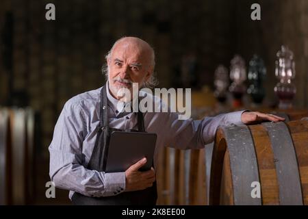 Caucasian senior man, a graybeard winemaker with apron checking big wooden barrels in a wine cellar and entering data in a tablet device Stock Photo