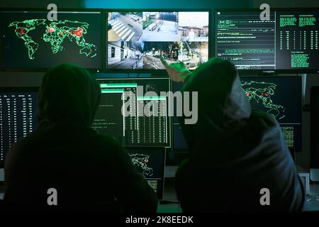 Back view of unrecognizable Criminal team Hacker pointing security cameras on screen monitor using computer for organizing massive data breach attack Stock Photo