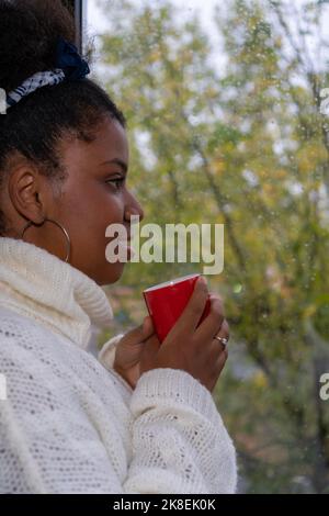 Young African American woman drinking coffee looking out the window on a rainy autumn day. Stock Photo