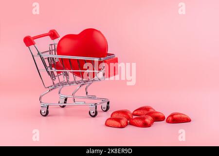 Red heart in shopping trolley and candies in a red wrapper isolated on pink background. Shopping for wedding, womens day, buying gifts Valentines Day. Stock Photo
