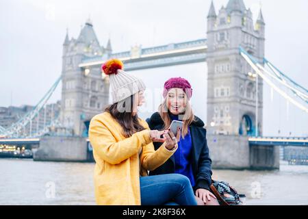 London, United KingdomTwo girls using smartphone in front of Tower Bridge Stock Photo