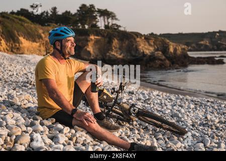 Thoughtful mature man sitting with bicycle on pebbles Stock Photo