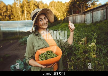 Smiling woman with bowl of green peas gesturing thumbs up in garden Stock Photo