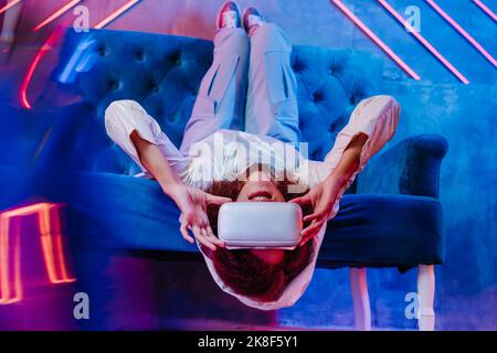 Woman with VR glasses lying upside down on sofa in front of neon light Stock Photo