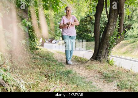 Active senior woman wearing wireless headphones walking by tree in park Stock Photo