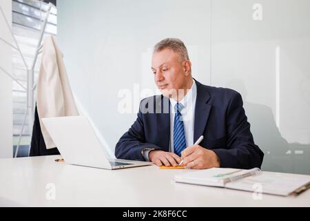 Mature businessman using laptop writing in sticky note at desk in office Stock Photo