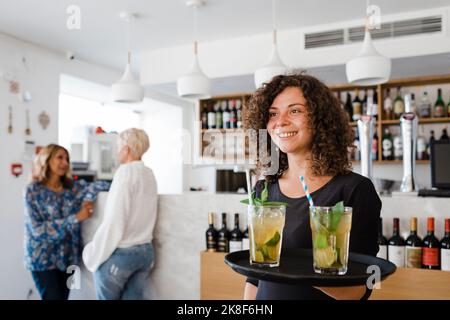 Happy young waitress with tray serving cocktail drinks at restaurant Stock Photo