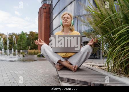 Mature woman with eyes closed sitting with laptop listening to music through headphones meditating on bench Stock Photo