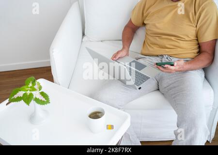 Freelancer with laptop using smart phone sitting in armchair at home Stock Photo