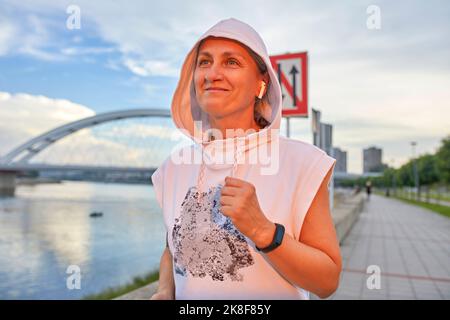 Smiling mature woman wearing in-ear wireless headphones jogging at promenade Stock Photo