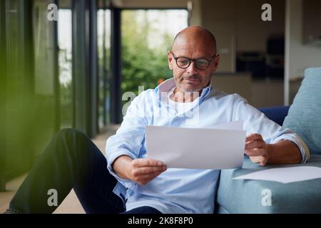 Senior man reading documents sitting by sofa at home Stock Photo