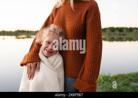 Smiling girl wrapped in blanket standing with mother at lake Stock Photo