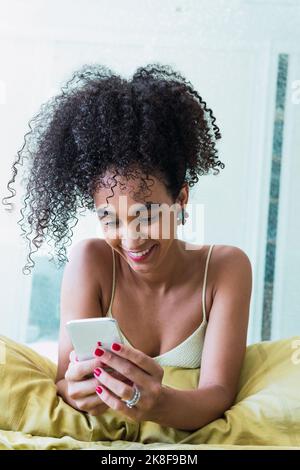 Happy curly-haired woman using smart phone at home Stock Photo