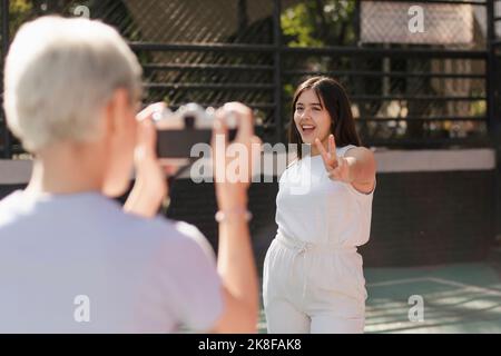 Happy woman gesturing peace sign in front of friend holding camera on sunny day Stock Photo