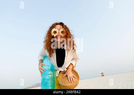 Smiling woman wearing sunflower sunglasses holding blue inflatable ring at beach Stock Photo