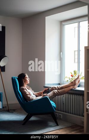 Thoughtful businesswoman sitting on chair with feet up at home Stock Photo