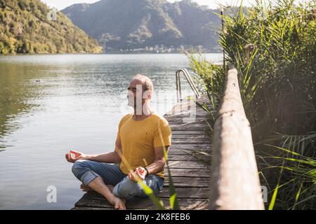 Mature man practicing lotus position on jetty by lake Stock Photo
