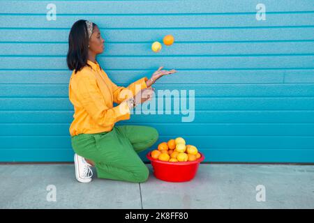Young woman juggling with oranges in front of blue roller shutter Stock Photo