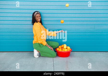 Young woman juggling with oranges in front of blue roller shutter Stock Photo