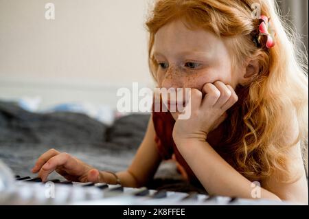 Girl with red hair playing synthesizer lying on bed Stock Photo