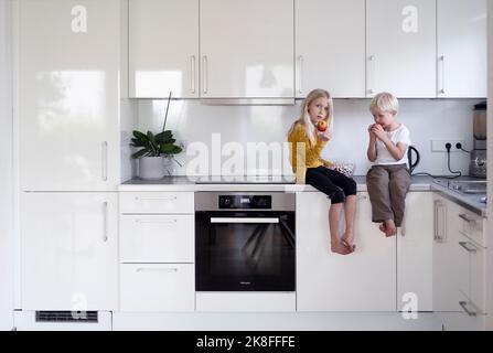 Boy and girl with apples sitting on kitchen counter at home Stock Photo