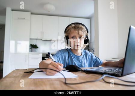 Thoughtful boy with wired headphones studying through laptop at home Stock Photo